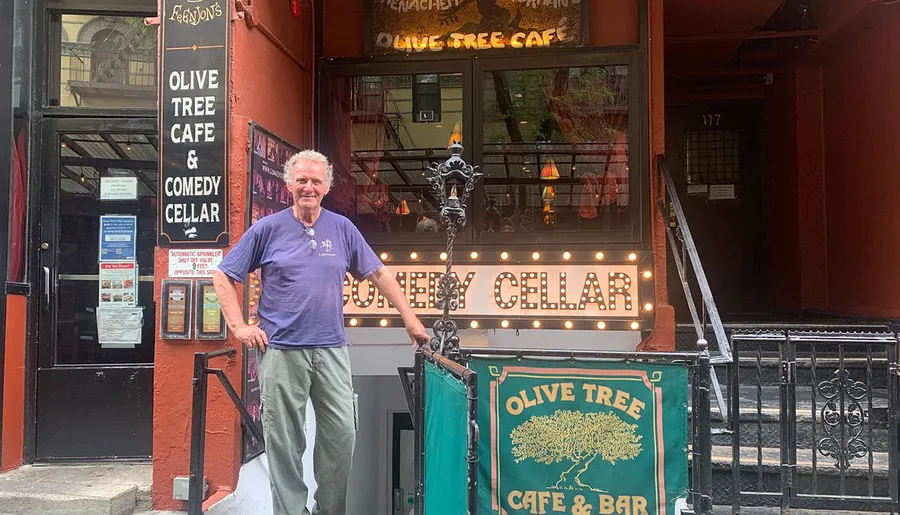 A man stands in front of the Olive Tree Cafe and Comedy Cellar, posing for the camera with a slight smile.