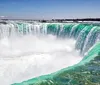 The image shows the powerful and picturesque Niagara Falls with cascades of water plunging over a crest lined with white frothy water and mist backed by a clear blue sky speckled with clouds