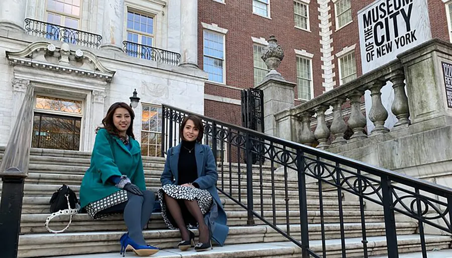 Two individuals are sitting on the steps outside the Museum of the City of New York.