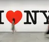 Two people are walking past the iconic I Love NY slogan displayed prominently on a gallery wall