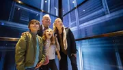 A family of four is looking up with expressions of wonder and amusement inside an elevator with illuminated glass walls.