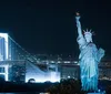 The image captures a nighttime scene of a Statue of Liberty replica with a brightly illuminated suspension bridge in the background