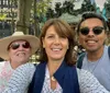 Three people are smiling for a selfie in front of a carousel in a bustling outdoor area dense with trees and street furniture