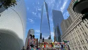 The image showcases a vibrant urban scene with the iconic One World Trade Center towering in the background, adjacent to distinctive architecture and a colorful mural, under a clear blue sky.