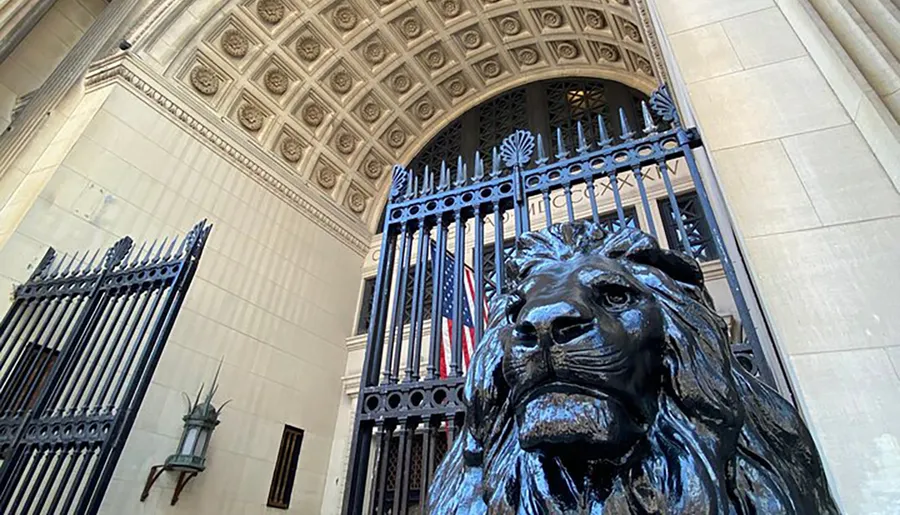 The image shows a majestic bronze lion statue foregrounding ornate gates with an American flag, part of an imposing architectural entrance.