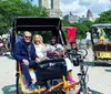 Three people are sitting in a pedal-powered rickshaw enjoying a sunny day in a park with other rickshaws and visitors in the background