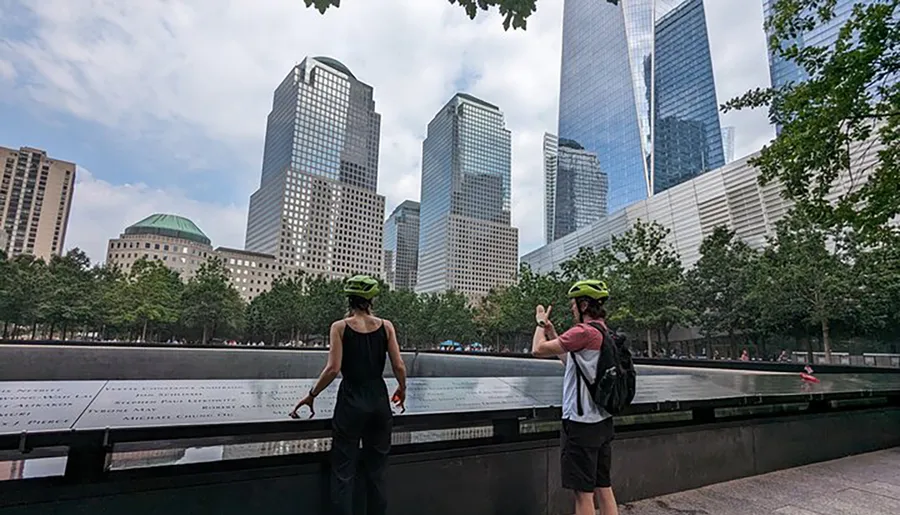 Two individuals with helmets are standing in front of a reflecting pool, with one person apparently making a peace sign, amid a backdrop of modern skyscrapers.