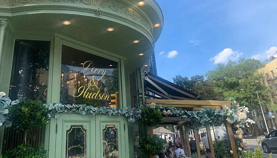 The image shows an elegant café exterior with a decorative façade and outdoor seating where patrons are enjoying their time under a clear blue sky.