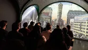 A group of people is watching a projection of a historic cityscape inside a room with arched openings.