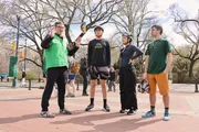 Four individuals are standing in a park-like setting, with the person on the left appearing to speak and gesture to the others who are attentively listening.