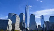 The image shows the Lower Manhattan skyline with One World Trade Center standing tall against a clear blue sky.