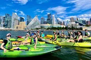 A group of kayakers enjoys a sunny day on the water with a backdrop of modern skyscrapers.