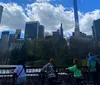 The image shows a view of Central Park with lush greenery in the foreground and a skyline of mixed architectural styles under a partly cloudy sky