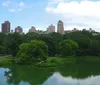 The image shows a view of Central Park with lush greenery in the foreground and a skyline of mixed architectural styles under a partly cloudy sky