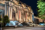 The image shows a grand, illuminated neoclassical building at night, possibly a museum, with a lit fountain in the foreground and people sitting on its steps.