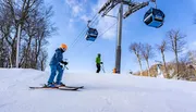 Two skiers are descending a snowy slope with a ski lift operating in the background under a clear blue sky.