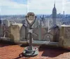 A coin-operated binocular viewer stands on an observation deck with a clear view of a city skyline featuring a prominent skyscraper under a blue sky