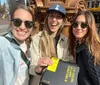 Three smiling individuals are taking a selfie on a sunny day on a city street holding a black binder or folder with a bright yellow label