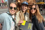 Three smiling individuals are taking a selfie on a sunny day on a city street, holding a black binder or folder with a bright yellow label.