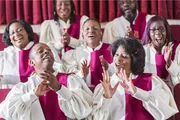 A joyful choir wearing white and red robes is singing and clapping with expressive and happy faces.