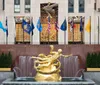 The image shows the iconic golden Prometheus statue overlooking the ice skating rink at Rockefeller Center against a backdrop of international flags and the bold artwork above the rink