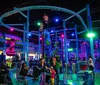 The image depicts an indoor amusement center with vibrant lighting featuring a roller coaster track above various play structures and people seated and walking around