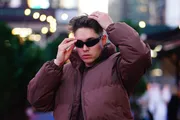 A person is adjusting their sunglasses and hair while wearing a brown jacket in an urban outdoor setting.