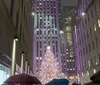 A vibrant Christmas tree illuminates a plaza surrounded by skyscrapers and people with umbrellas on a rainy evening