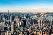 An aerial view showcases a dense array of skyscrapers and buildings in a bustling metropolis, with a wide river visible in the background.