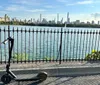 An electric scooter stands abandoned by an ornate metal fence with a scenic view of a city skyline across a body of water