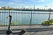 An electric scooter stands abandoned by an ornate metal fence with a scenic view of a city skyline across a body of water.