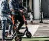 An electric scooter stands abandoned by an ornate metal fence with a scenic view of a city skyline across a body of water