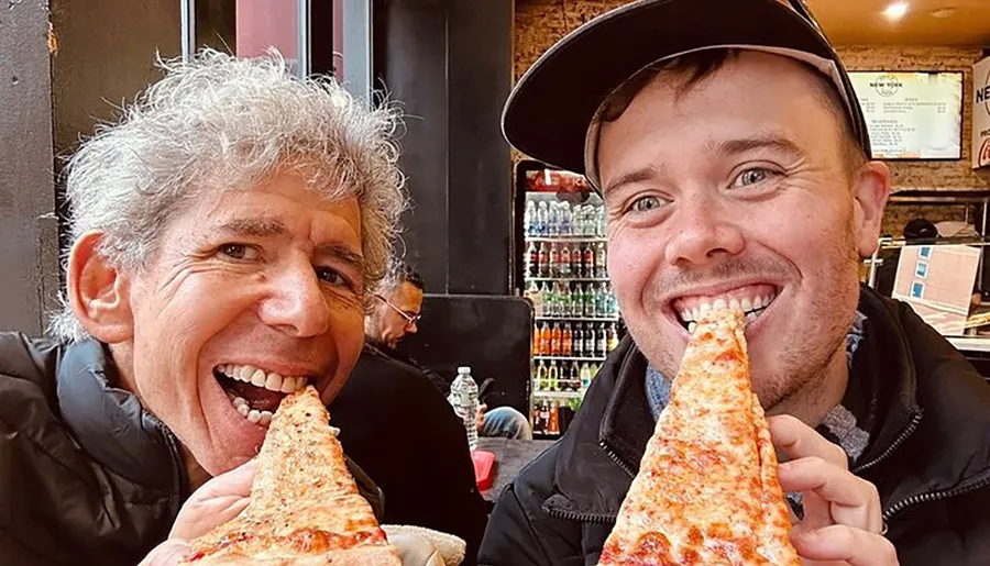 Two people are smiling at the camera while biting into large slices of pizza.