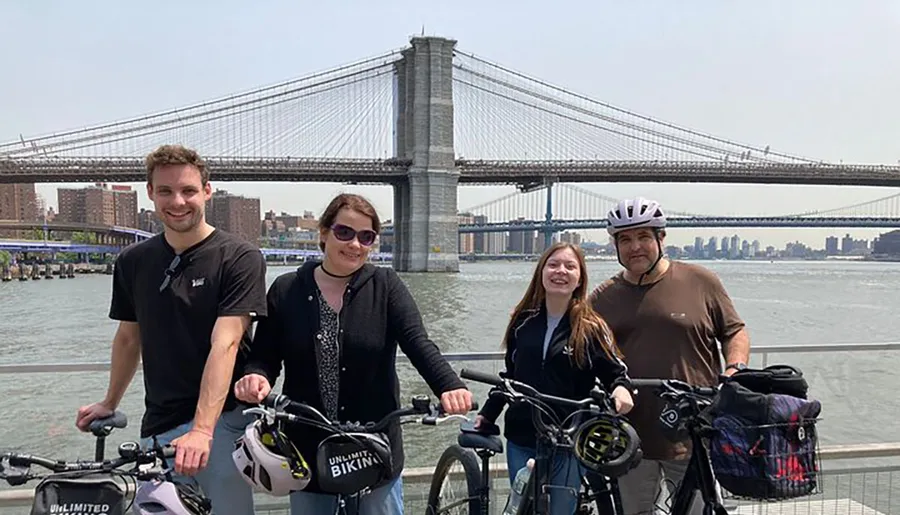 Four individuals pose with their bicycles on a waterfront with the Brooklyn Bridge in the background.