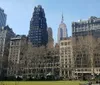 The image shows a view of a park in an urban setting with the Empire State Building towering in the background amidst other high-rise buildings under a clear blue sky