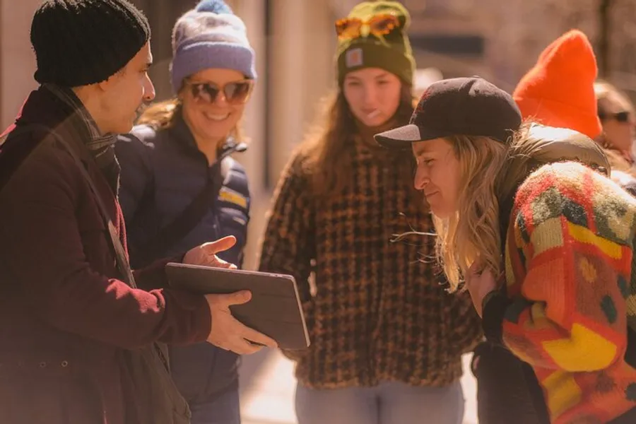 A group of people, some wearing colorful winter hats, gather around a person holding a tablet, engaged in what seems to be an interesting interaction or discussion in an outdoor setting.