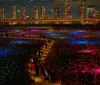 The image shows a vibrant field of multicolored lights in the foreground with visitors walking through set against a backdrop of a city skyline at night