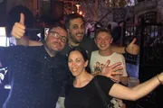 Four friends are posing for a group photo with smiles and thumbs-up gestures, seemingly enjoying a good time together in a bar-like setting.