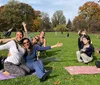 A group of people are posing joyfully on yoga mats in a sunny park with autumn foliage in the background