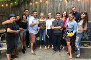 A group of smiling people are posing for a photo at an outdoor gathering, some holding drinks and snacks, suggesting a social event or celebration.