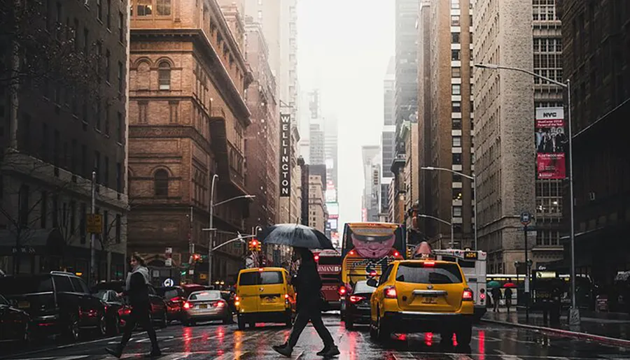 Pedestrians with umbrellas and yellow cabs navigate through a rain-soaked street in a bustling city.