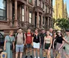 A group of smiling people poses for a photo on the sidewalk in front of a row of brownstone buildings indicative of an urban residential neighborhood