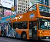 An orange double-decker sightseeing tour bus is navigating through an urban street prominently advertising Hop On Hop Off tours with city imagery