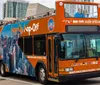 An orange double-decker sightseeing tour bus is navigating through an urban street prominently advertising Hop On Hop Off tours with city imagery