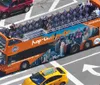 An orange double-decker sightseeing tour bus is navigating through an urban street prominently advertising Hop On Hop Off tours with city imagery