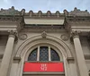 The image shows the classical facade of The Metropolitan Museum of Art in New York with a prominent red banner featuring the museums abbreviated name THE MET