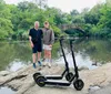 Two individuals and two electric scooters are standing on a rock with a pond and an arched bridge in the background