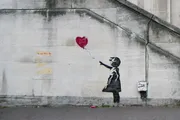 The image shows a stencil graffiti by the artist Banksy, depicting a young girl reaching out toward a red, heart-shaped balloon.