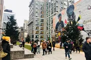 People stroll by a modern building with an artistic mural, near a Christmas tree, in an urban park setting.