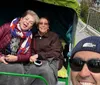 Three people are smiling for a selfie with the person in the foreground giving a thumbs up and the two in the background seated on a park bench waving at the camera